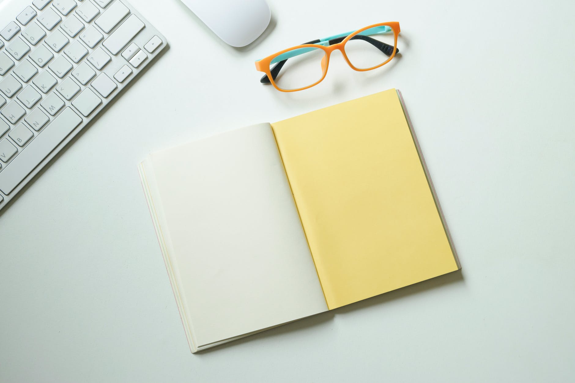 white and yellow notebook placed near keyboard and eyeglasses